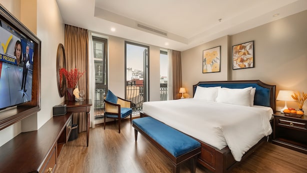 A hotel room with a large bed, a desk, a chair, a bench, a TV, and a balcony with a view of the city. - Hanoi Fiesta Grand Hotel (Hanoi)
