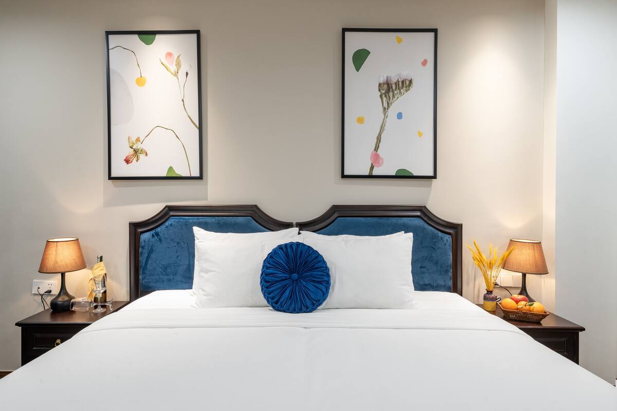 A neatly made bed with a blue accent pillow, flanked by two bedside tables with lamps and a fruit basket.