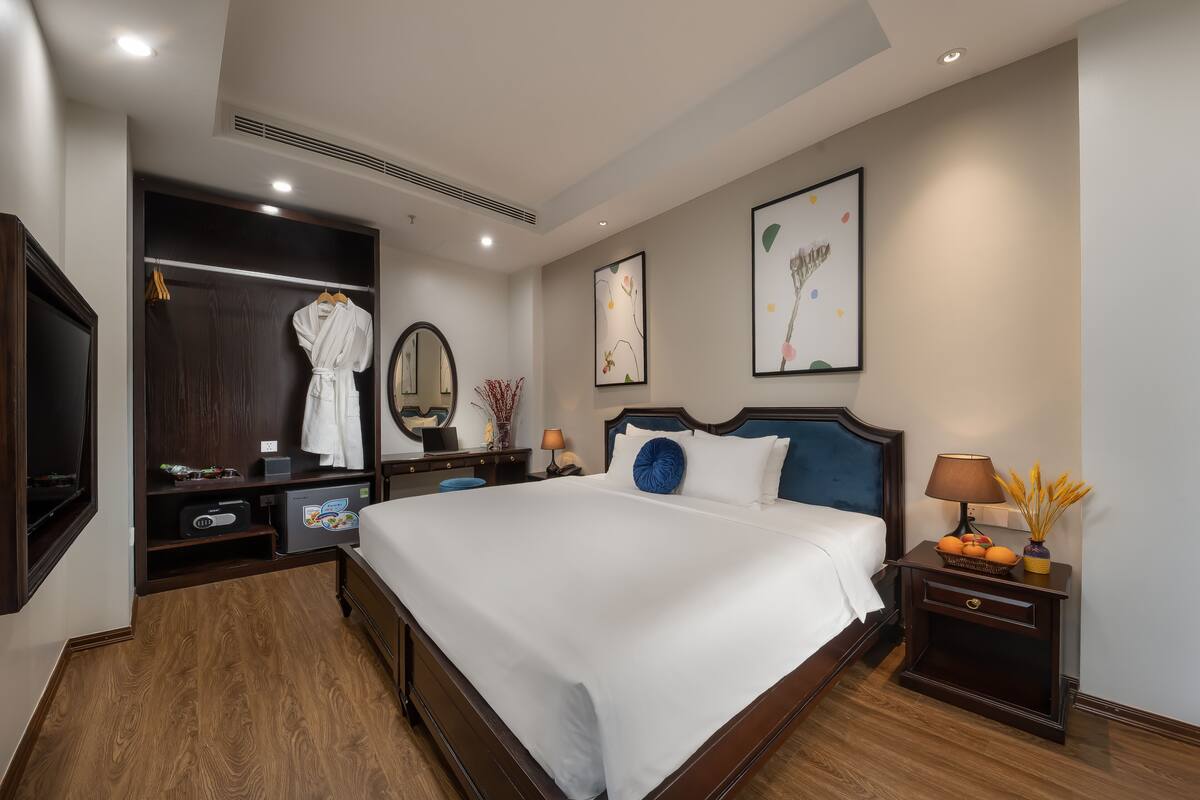 A hotel room with a large bed, a wardrobe, a bedside table with a lamp, and artwork on the walls.