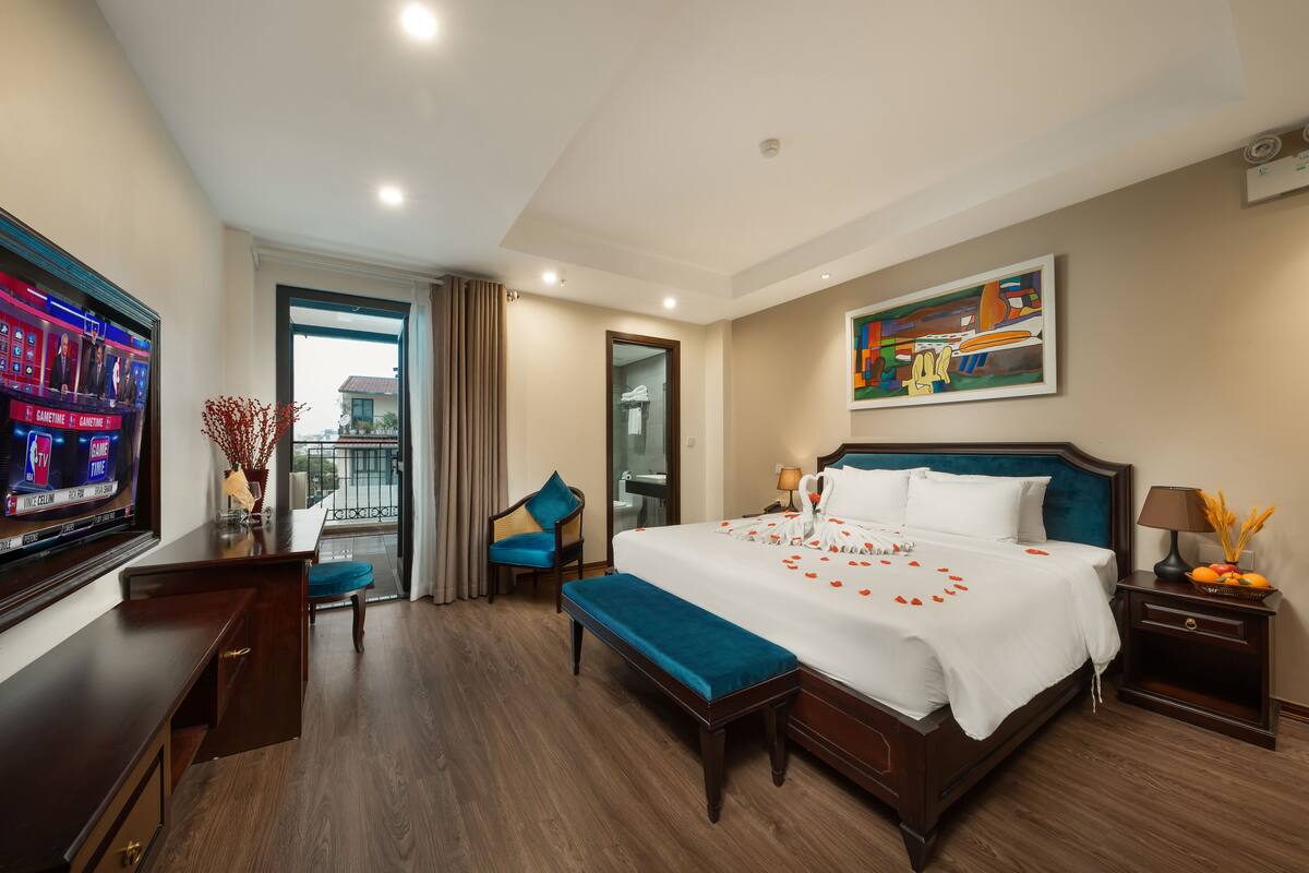 A hotel room with a large bed, a TV, a desk with a chair, a bench, and a nightstand with a lamp.