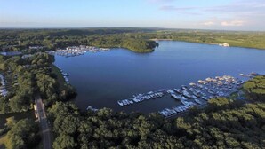 Luftaufnahme einer Marina, umgeben von dichtem Wald, mit vertäuten Booten und blauem Himmel.