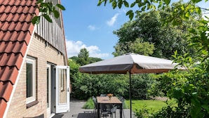Eine Terrasse mit Tisch und Stühlen unter einem großen Sonnenschirm, ein Ziegelhaus mit rotem Dach und üppiges Grün im Hintergrund.