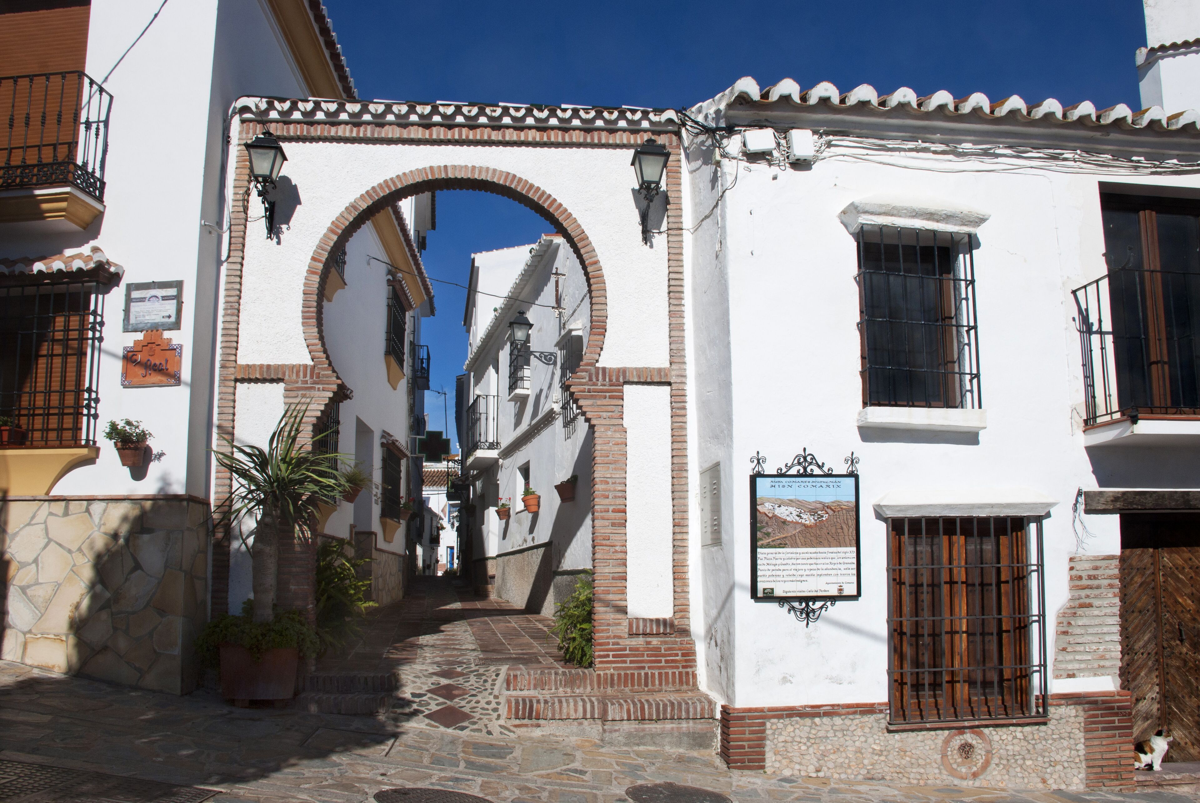 A narrow street with white buildings, a stone-paved path, and a large arched doorway.