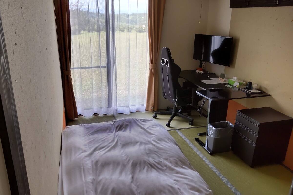 A hotel room with a bed, a desk with a computer, a chair, a TV, and a window with curtains.