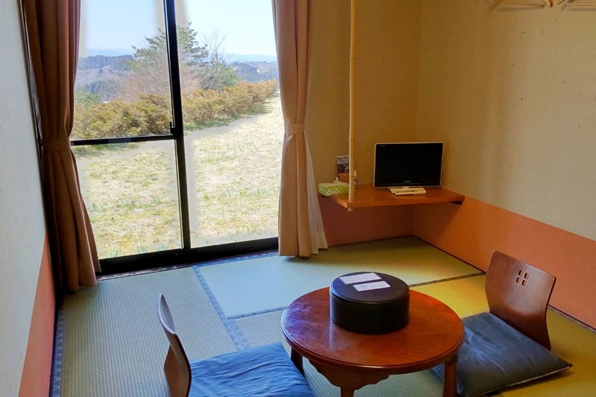 A room with tatami mat flooring, a low table with a cushion, a chair, a TV on a shelf, and a view of a natural landscape through a large window.