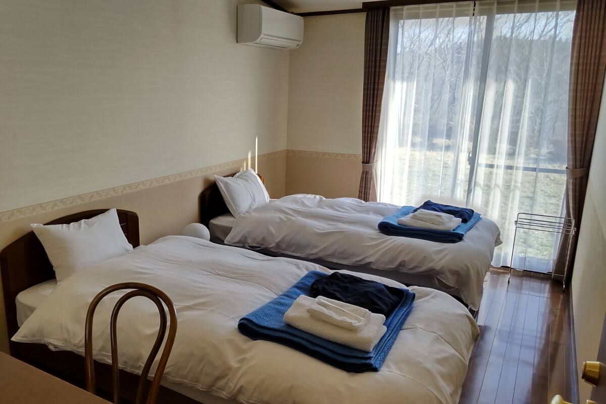 A hotel room with two beds, a wooden chair, a window with curtains, and an air conditioning unit.