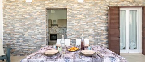 A dining table set with plates, cutlery, and bottles, against a stone wall with a kitchen visible through an open door.