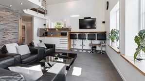 Een moderne woonkamer met een zwarte leren bank, een glazen salontafel en een keukenhoek met een televisie en barstoelen.