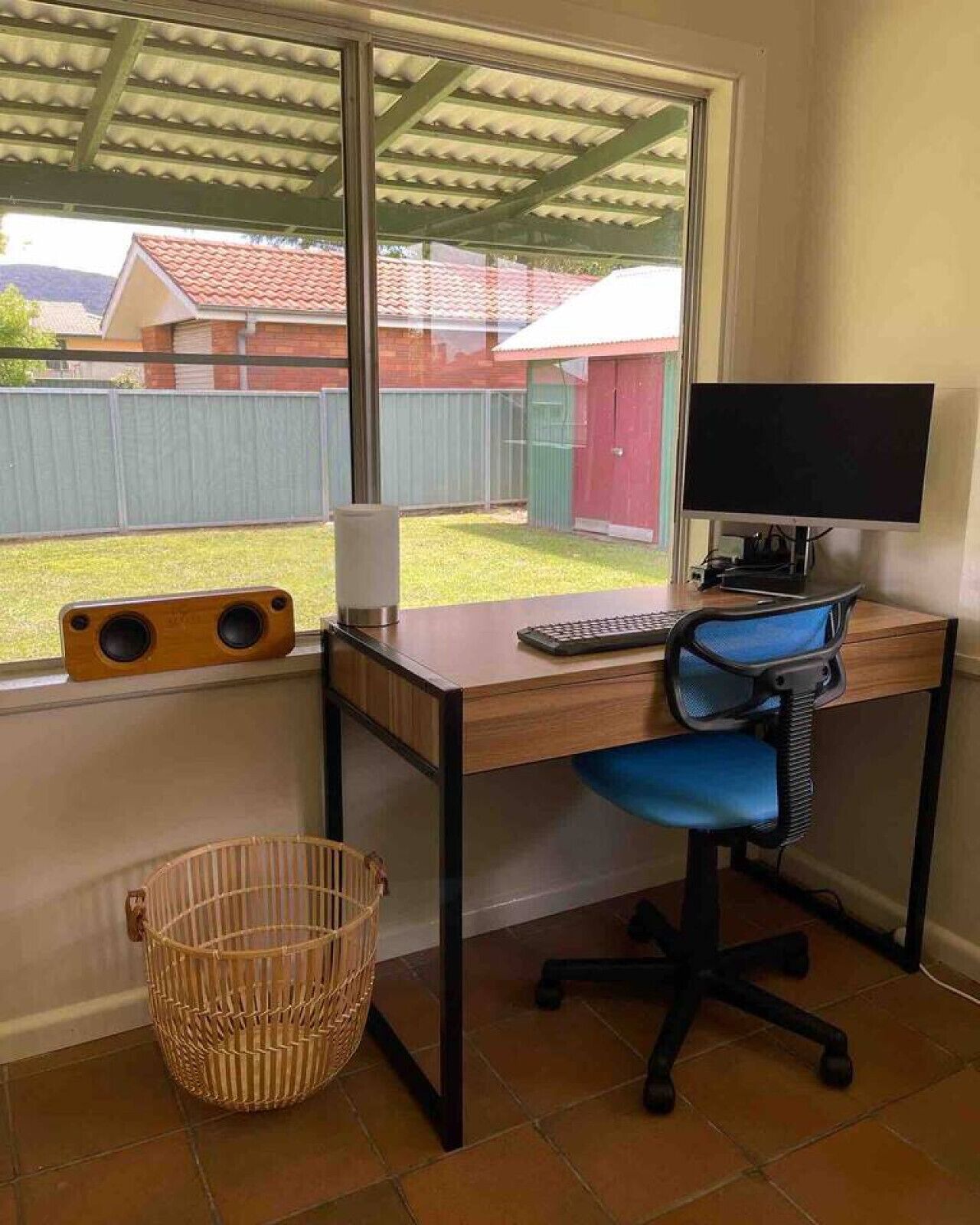 A home office with a desk, computer, and chair, a window with a view of a backyard, and a wicker basket.