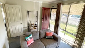 A living room with a grey sofa, two colorful cushions, a metal rack with bottles, and a view of a backyard through a sliding door. - Comfortable 2-Bedroom home “Rest Easy Mudgee” (Mudgee)