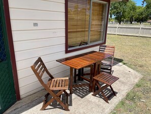 A wooden table and chairs set outside a building with a window and a white picket fence in the background.