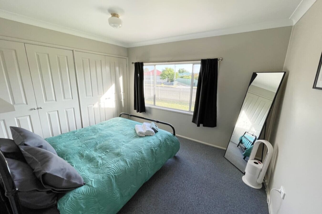 A bedroom with a bed, a window with curtains, a mirror, and a white wardrobe.