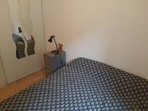 Quarto com colcha estampada, cômoda com gavetas, luminária e espelho com design ondulado na parede.