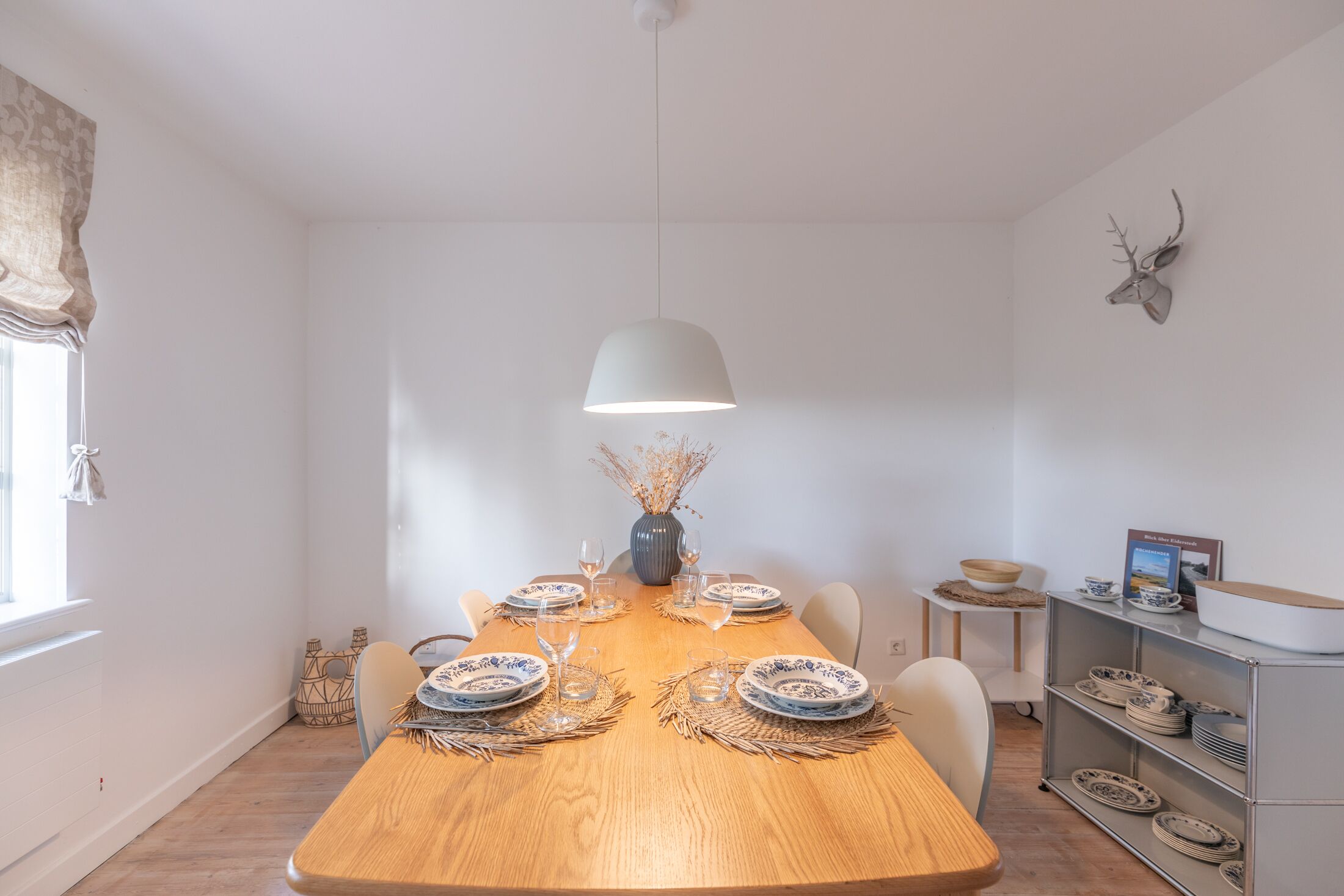 A dining area with a wooden table set for four, a hanging light fixture, and a wall-mounted deer head decoration.