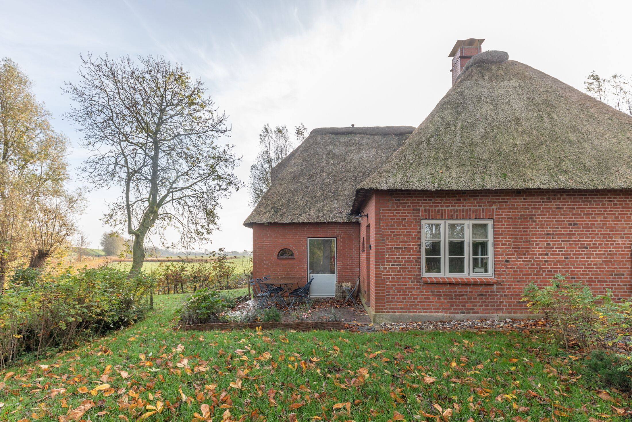 A brick house with a thatched roof, a small porch with chairs, and a garden with fallen leaves.