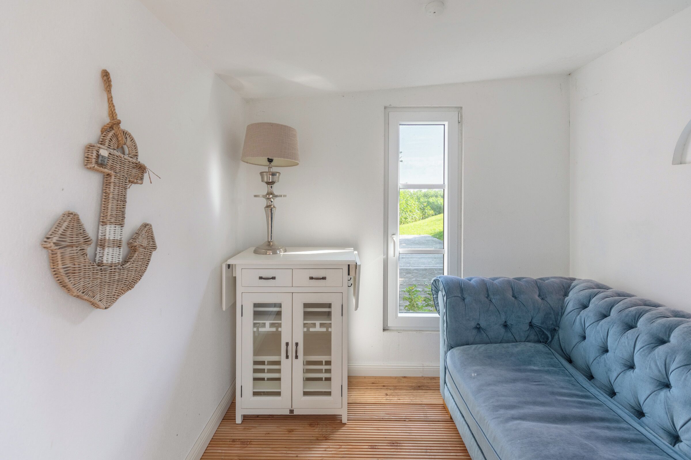 A room with a blue sofa, a white cabinet with glass doors, a lamp, and a view of greenery through a window.