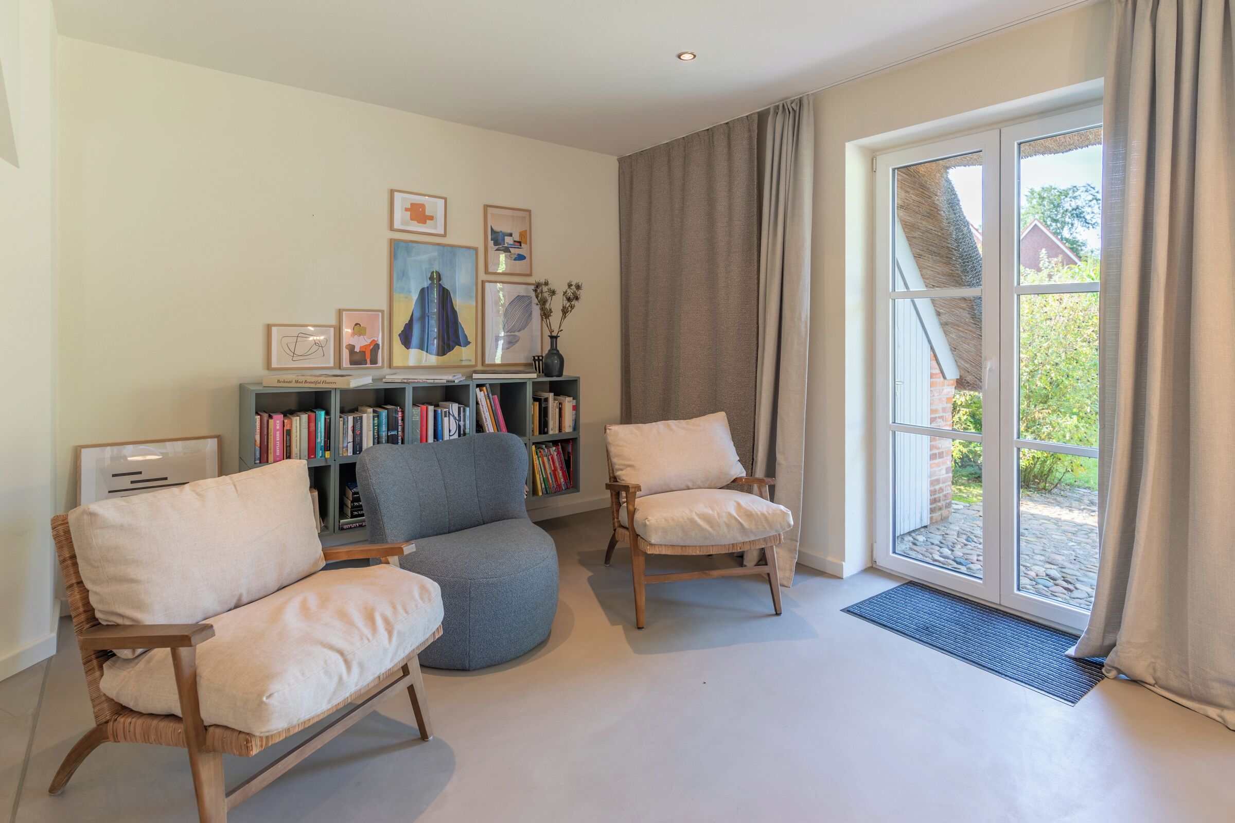 Un salon avec un canapé gris, deux fauteuils en bois, une bibliothèque et une grande fenêtre avec des rideaux.