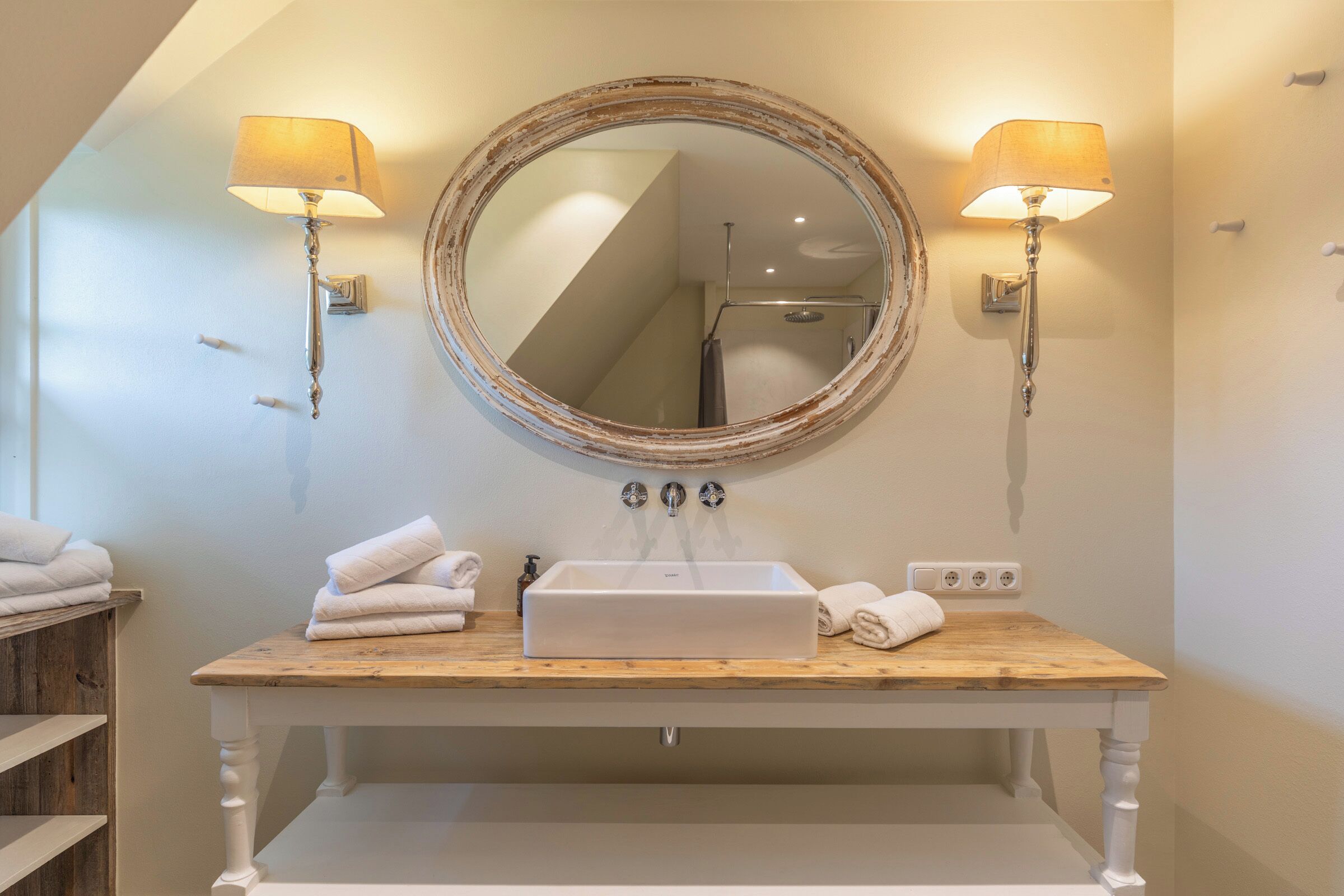 A bathroom with a wooden vanity, a large mirror, two wall-mounted lamps, and a shower visible in the background.