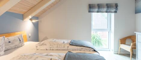 A bedroom with a sloped ceiling, a bed with patterned bedding, a window with a view of greenery, and a wicker chair.