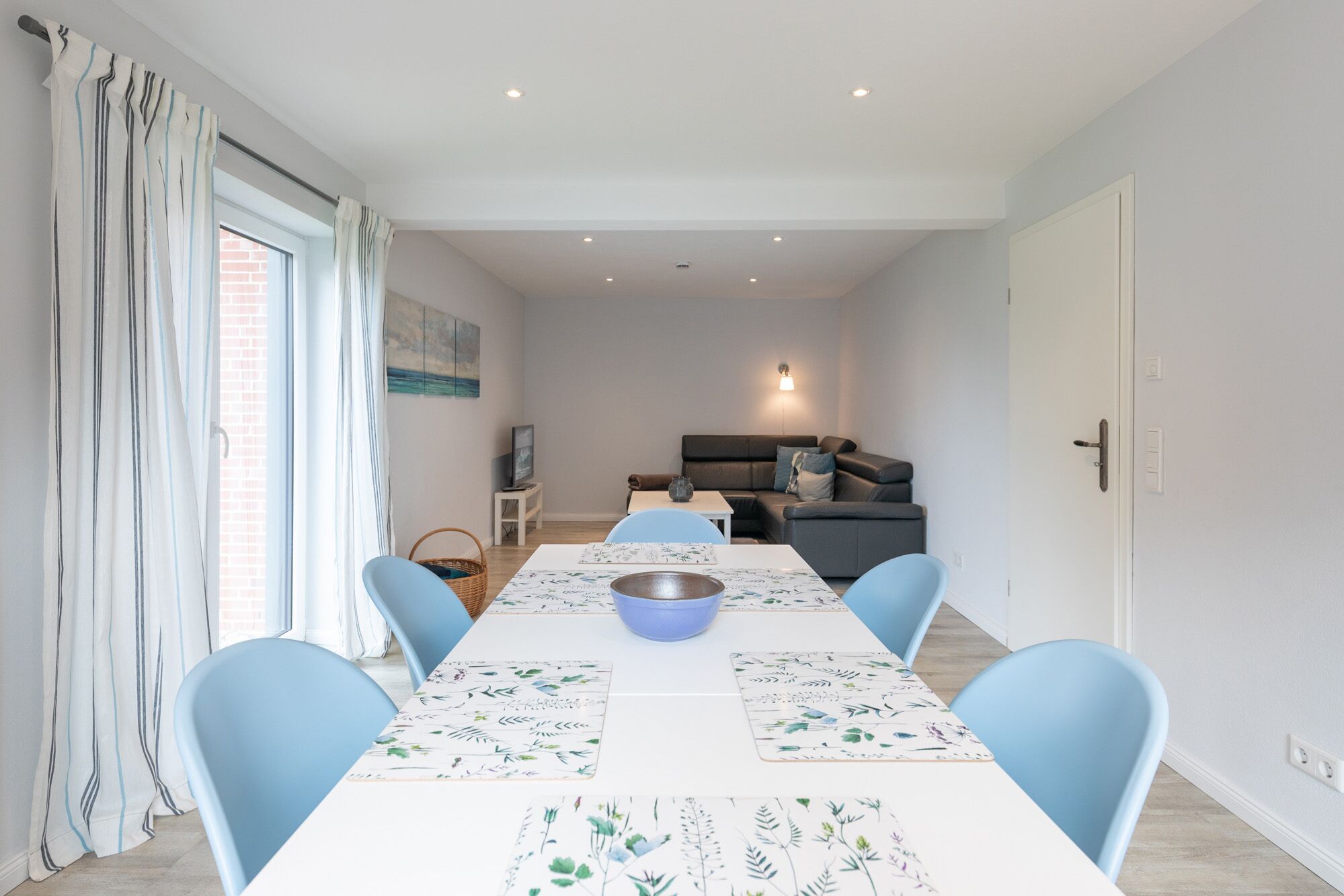 Un coin repas avec une longue table, des chaises bleues, un saladier et un téléviseur fixé au mur.