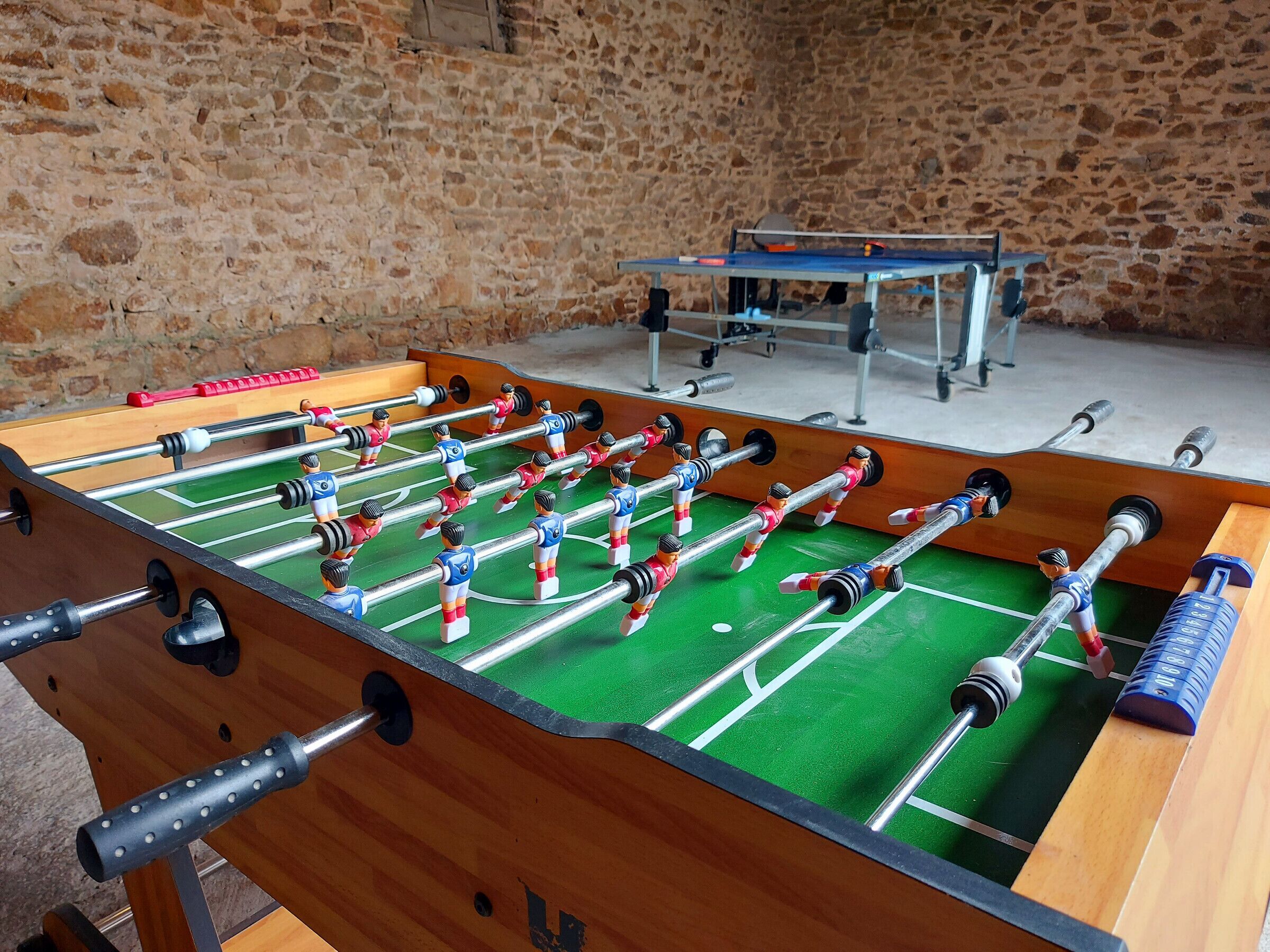 Een tafelvoetbaltafel met meerdere spelers en een tafeltennistafel in een ruimte met stenen muren.