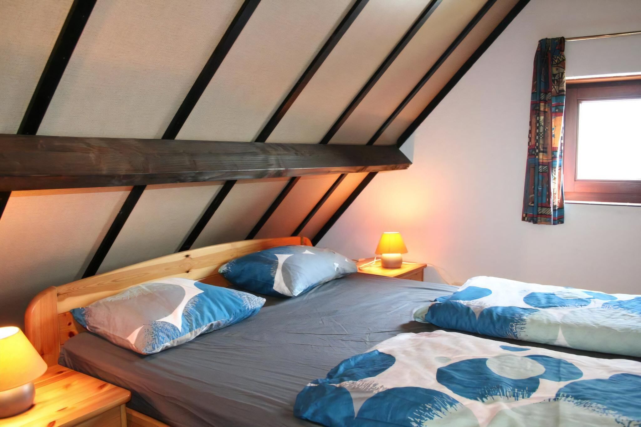 A bedroom with a sloped ceiling, a wooden headboard, a bed with blue and white bedding, a bedside lamp, and a window with patterned curtains.