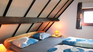 Quarto com teto inclinado, cabeceira de madeira, cama com roupa de cama azul e branca, luminária de cabeceira e janela com cortinas estampadas.