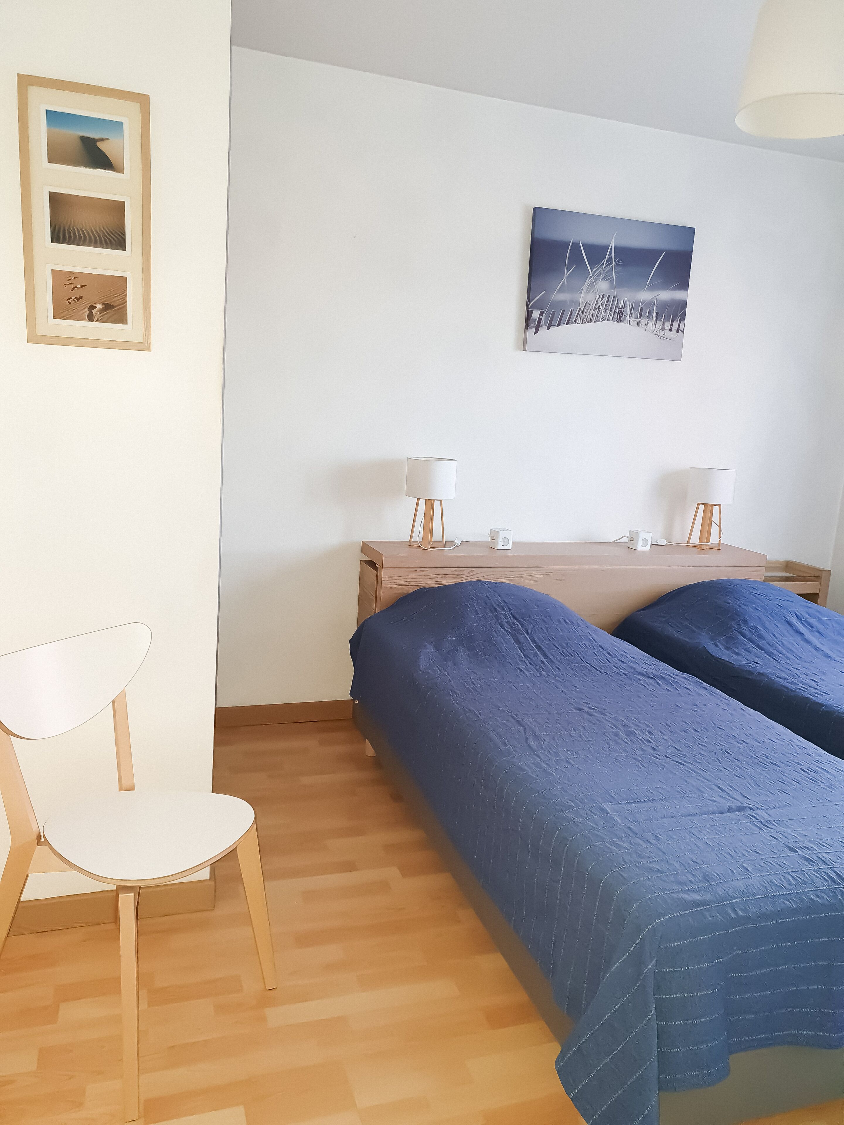 Un dormitorio con dos camas, una silla, una mesita de noche con lámparas y una pared decorada con cuadros.