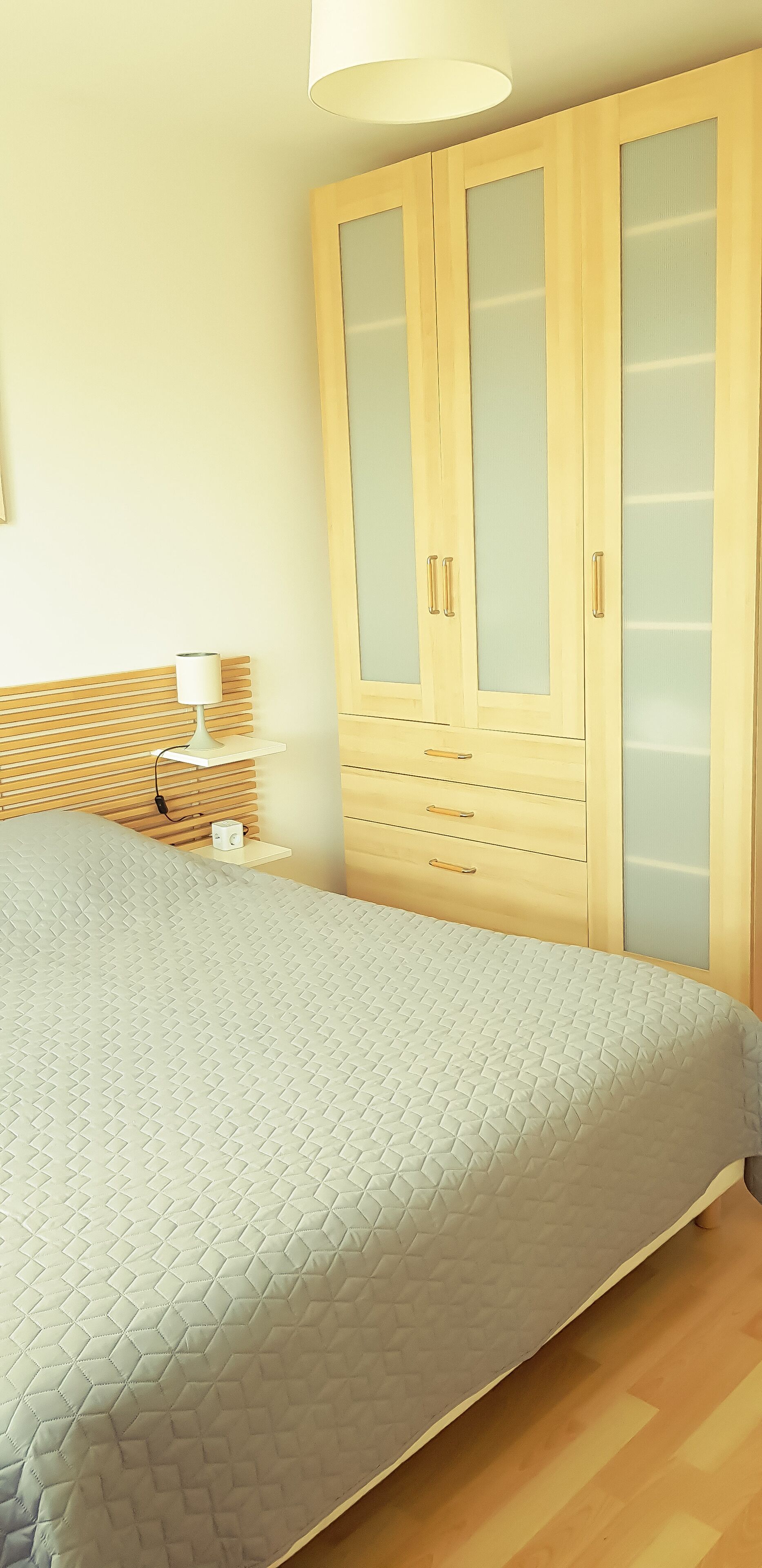 Une chambre à coucher comprenant un lit, une armoire en bois avec des portes en verre, une table de chevet avec une lampe et une étagère fixée au mur.