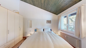 Un dormitorio con cama, armario, ventana con cortinas, radiador y un cuadro pequeño en la pared.