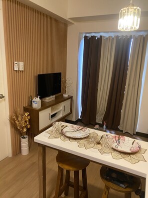 Une cuisine moderne avec un mur recouvert de panneaux en bois, un téléviseur à écran plat, une table à manger agrémentée d’un chemin de table décoratif et une fenêtre avec des rideaux.