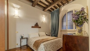 Un dormitori amb un llit de fusta, una tauleta de nit, un aparador, una finestra amb cortines i una làmpada fixa a la paret.