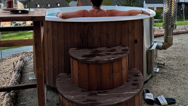 Eine Person entspannt sich in einer Badewanne mit Blick auf einen Sonnenuntergang und Häuser im Hintergrund.
