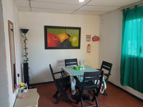 A dining area with a table set for four, chairs, a framed fruit painting, a fire extinguisher, and a green curtain. - Casa Camimar (Santa Marta)