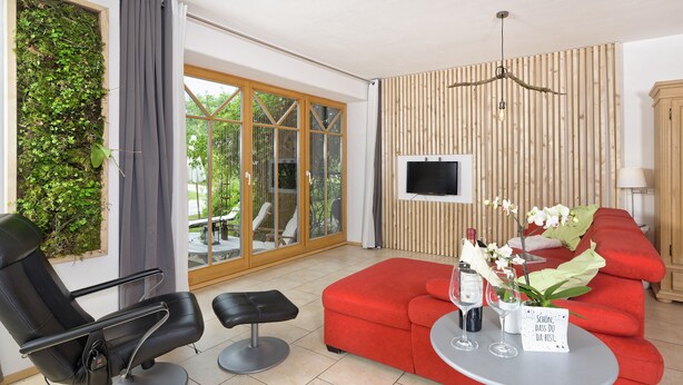 Een moderne woonkamer met een rode bank, een zwarte fauteuil, een klein tafeltje met drankjes en een televisie bevestigd aan een houten wand.