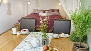 Een gezellige kamer met een bed, een eettafel met een tafelkleed met bloemenprint, een schaal appels, een theepot, een klein plantje en een potplant.