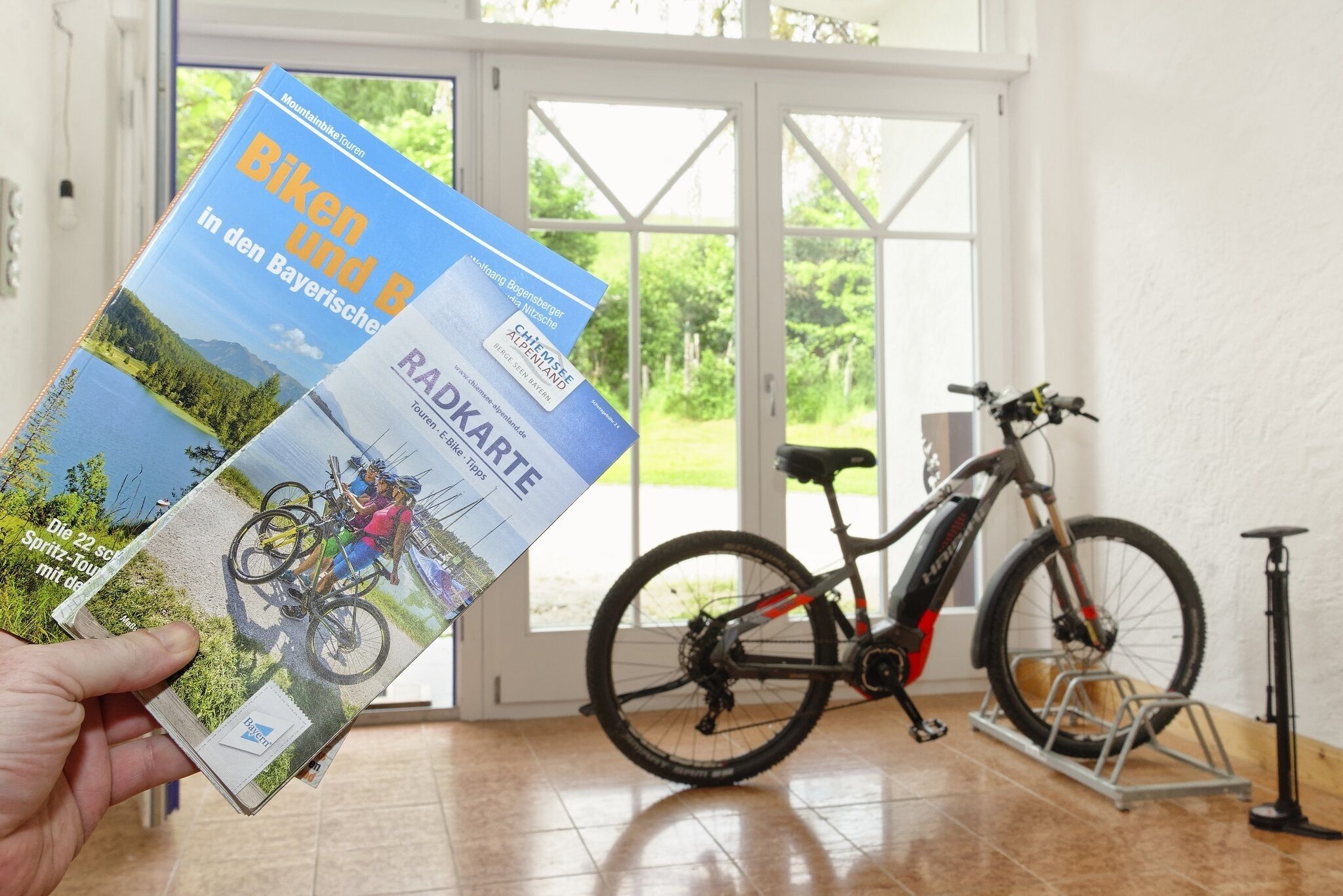 A person holding a brochure titled "Biken und Radkarte" in front of a bicycle and a bike rack.