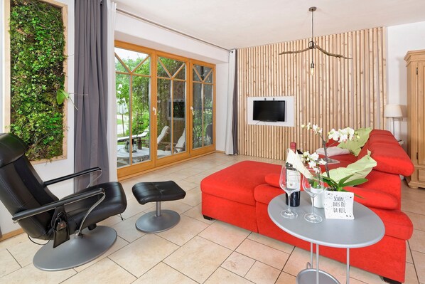 A modern living room with a red sofa, a black chair, a small table with a bottle and glasses, and a TV mounted on the wall.
