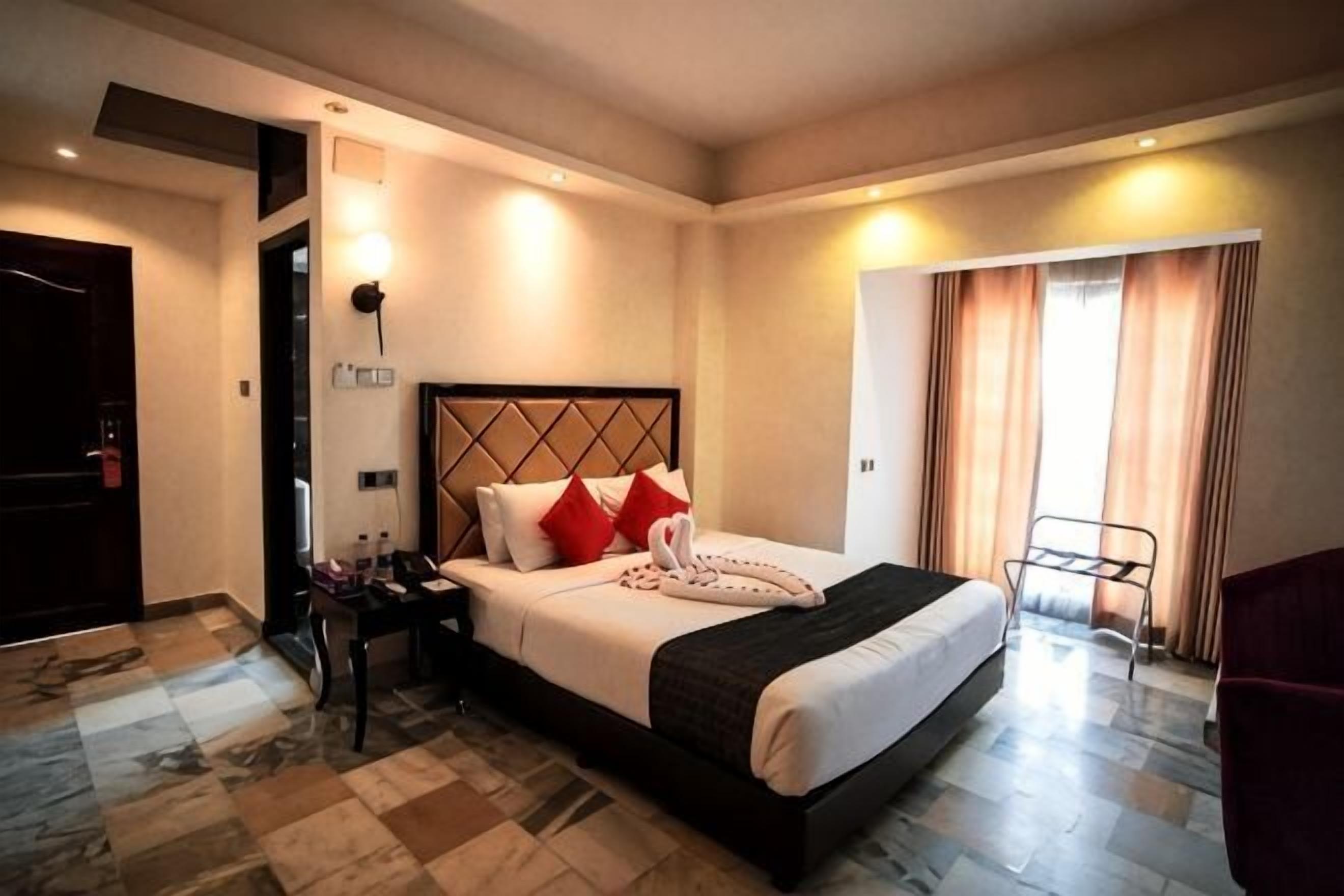 A hotel room with a bed, bedside table, lamp, and a view of the outside.