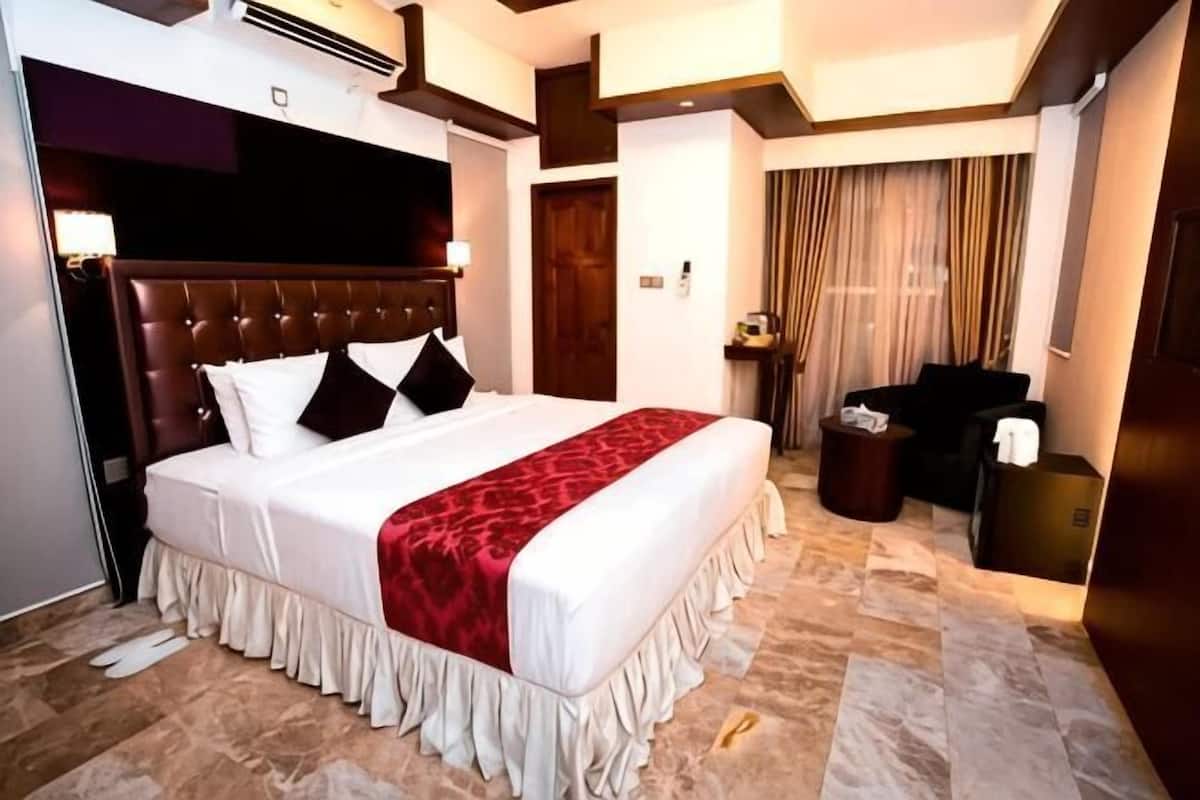 A hotel room with a large bed, a wooden headboard, a red throw blanket, a small round table, and a dark-colored sofa.