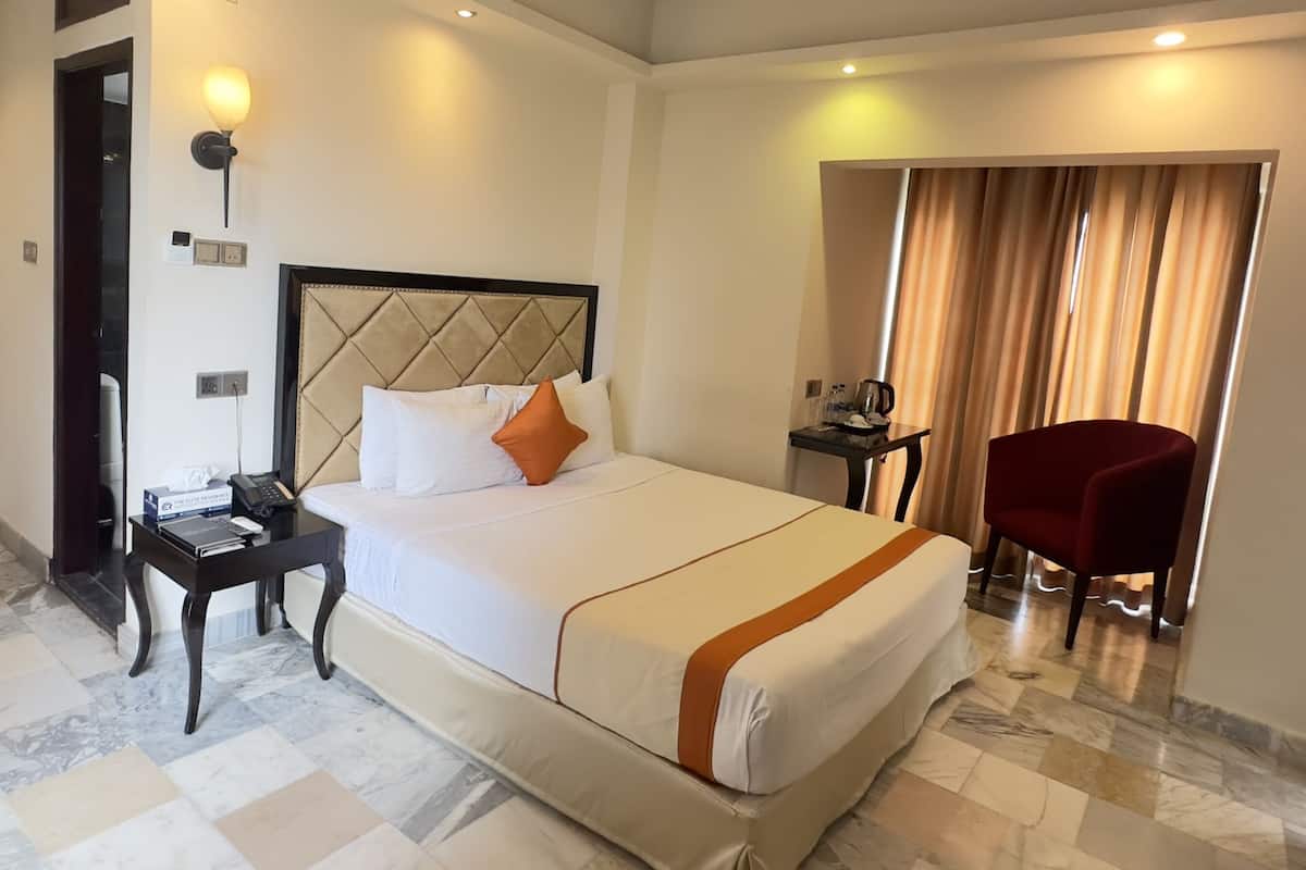 A hotel room with a large bed, a bedside table with a phone and a book, a red chair, and a small table with a kettle and cups.