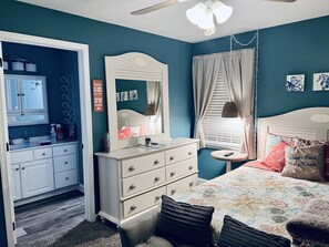 Quarto com cama, penteadeira, cômoda e uma pequena mesa redonda com abajur.