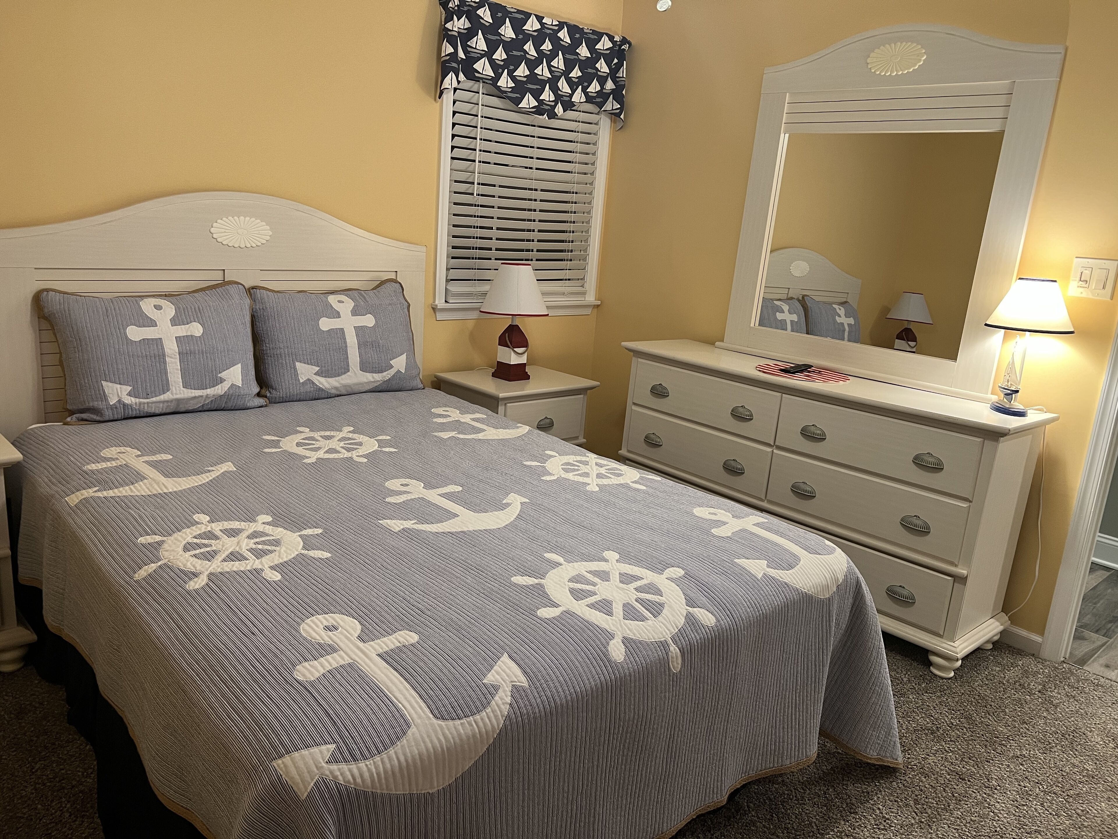 Un dormitorio con una cama que tiene una colcha con motivos náuticos, una mesita de noche con lámpara, un aparador con espejo y una ventana con cortina estampada.