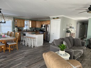Un salón amplio con un sofá gris, un reposapiés de cuero color beige y una mesa de comedor con sillas de madera. Se observa una cocina en el fondo, con armarios de madera y una isla blanca.