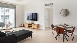 A modern living room with a black sofa, a wooden cabinet, a flat-screen TV, and a dining area with a round table and chairs. - Luxury 3BR 32 Floor SeaView (Bat Yam)