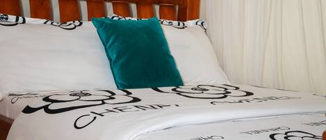 A bed with a floral bedspread and pillows, a wooden headboard, and a white curtain in the background.