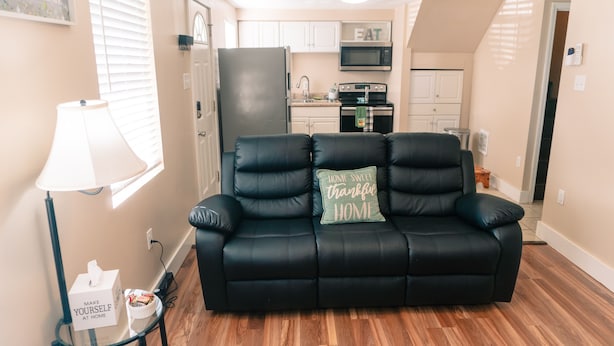 Una sala de estar con un sofá de cuero negro, una lámpara, una mesita con una caja de pañuelos y un cartel que dice "MAKE YOURSELF AT HOME".
