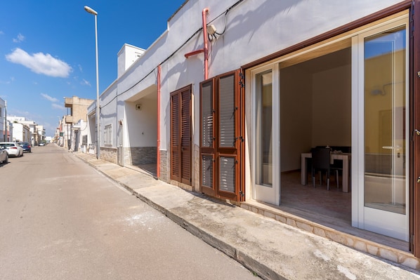 A street view of a building with a covered outdoor seating area and a visible interior space with a table and chairs.