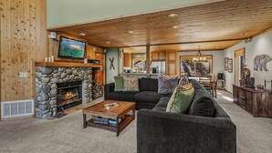 A cozy living room with a stone fireplace, a television, and a large window overlooking a scenic view. - Martis Valley Vistas: Family Home w/ Private Hot Tub, 2 Living Rooms, near Golf (Truckee)