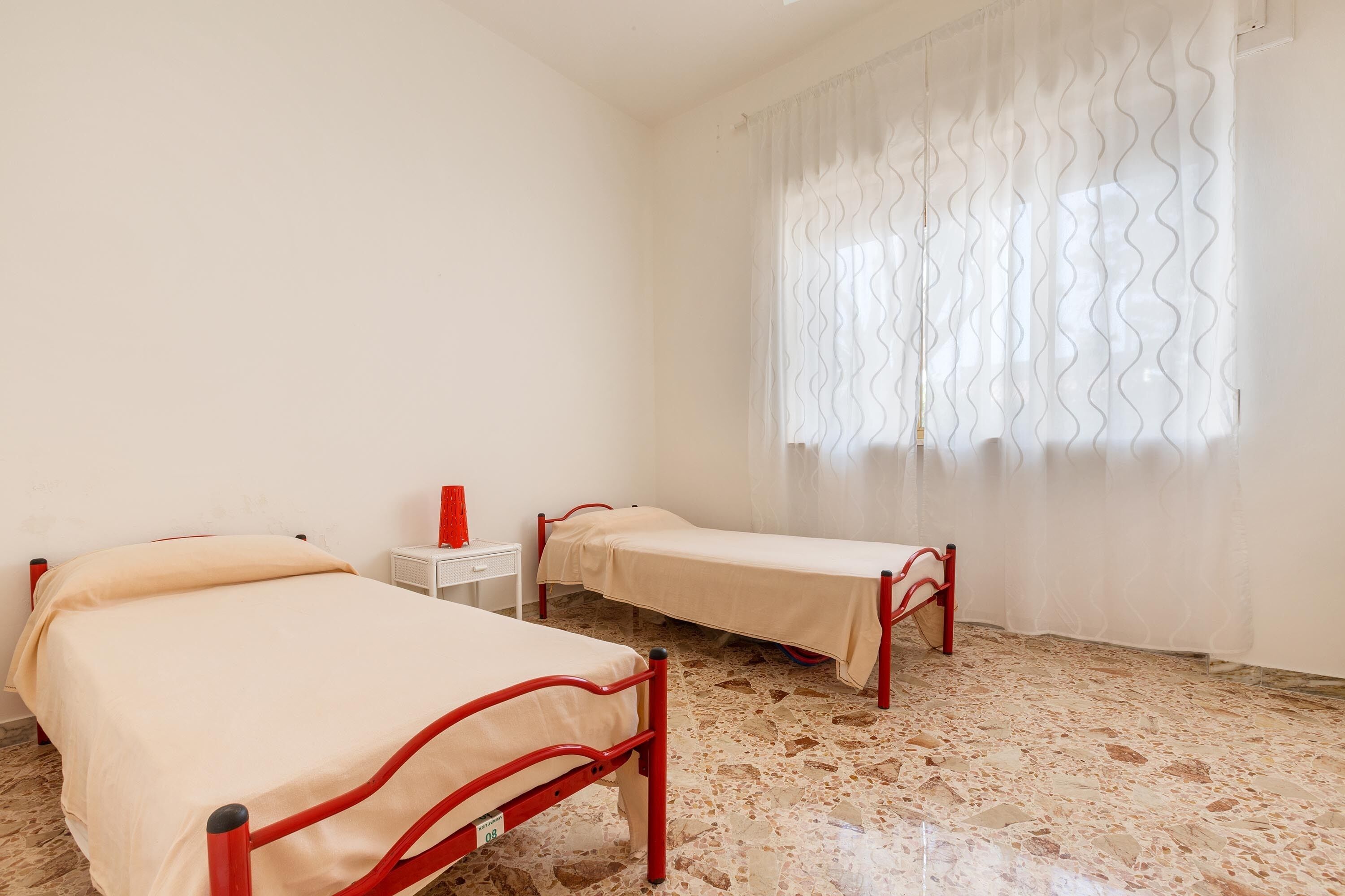 A room with two single beds, a small bedside table, and a window with sheer curtains.